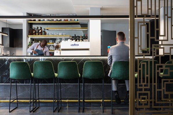 Een bar met een bartender die drankjes bereidt, een toonbank met verschillende flessen en groene barkrukken.