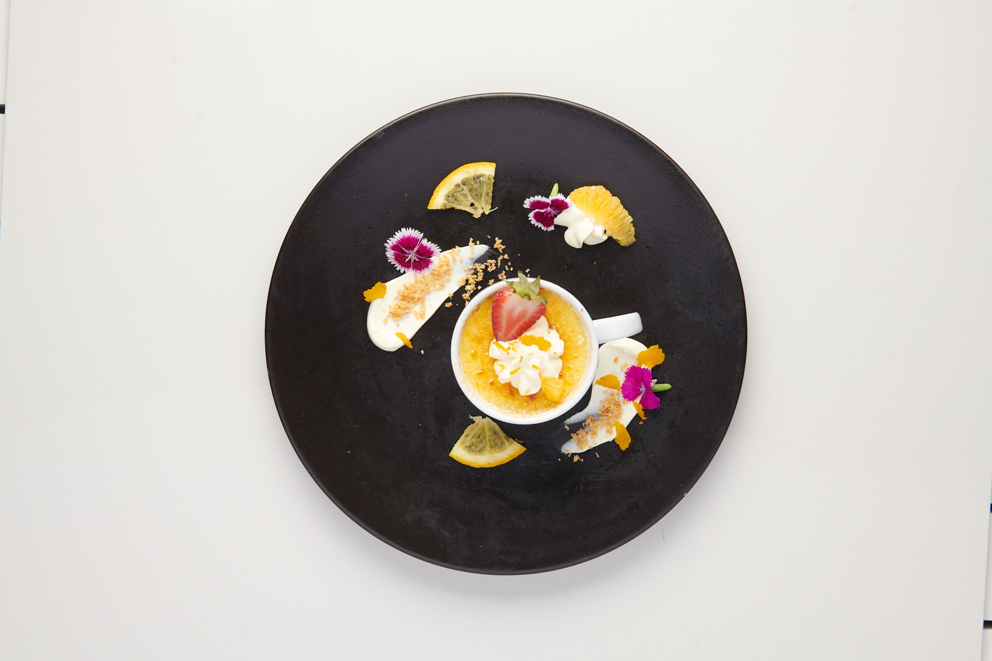 Un plato negro redondo con un postre en el centro, rodeado de diversas frutas y flores.