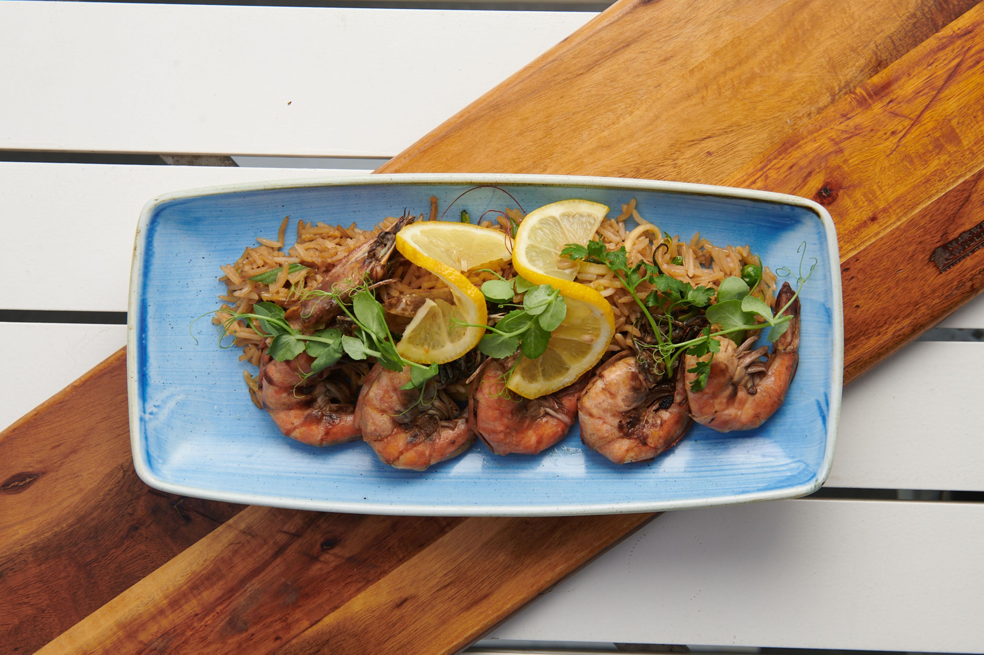 Une assiette bleue contenant des crevettes grillées, des tranches de citron et des légumes verts, le tout disposé sur une planche en bois.