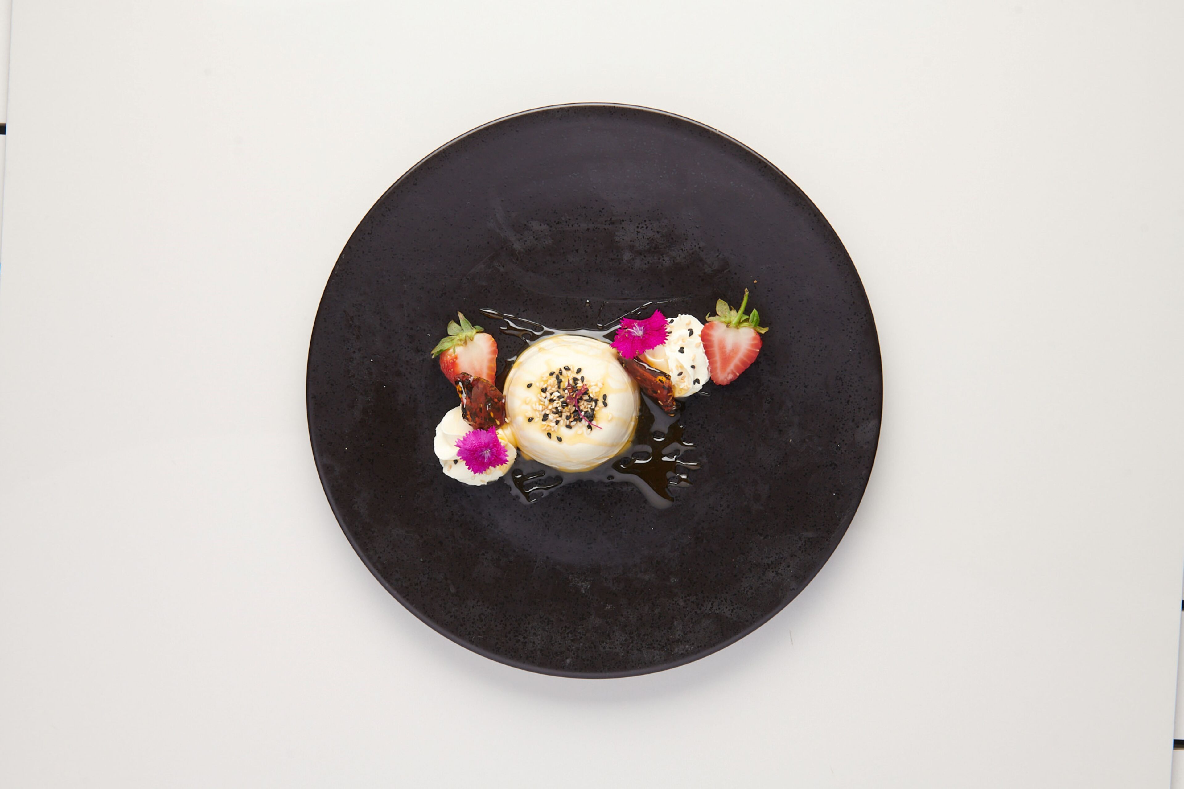 Un plato negro redondo con un postre de fresas y una flor.