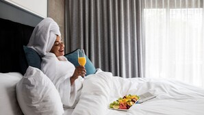 Een persoon in een witte badjas zittend op een bed met een schaal fruit en een glas sinaasappelsap.