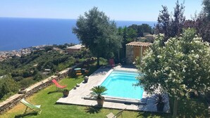 Uma piscina com deck de pedra, rodeada por vegetação e vista para o mar.