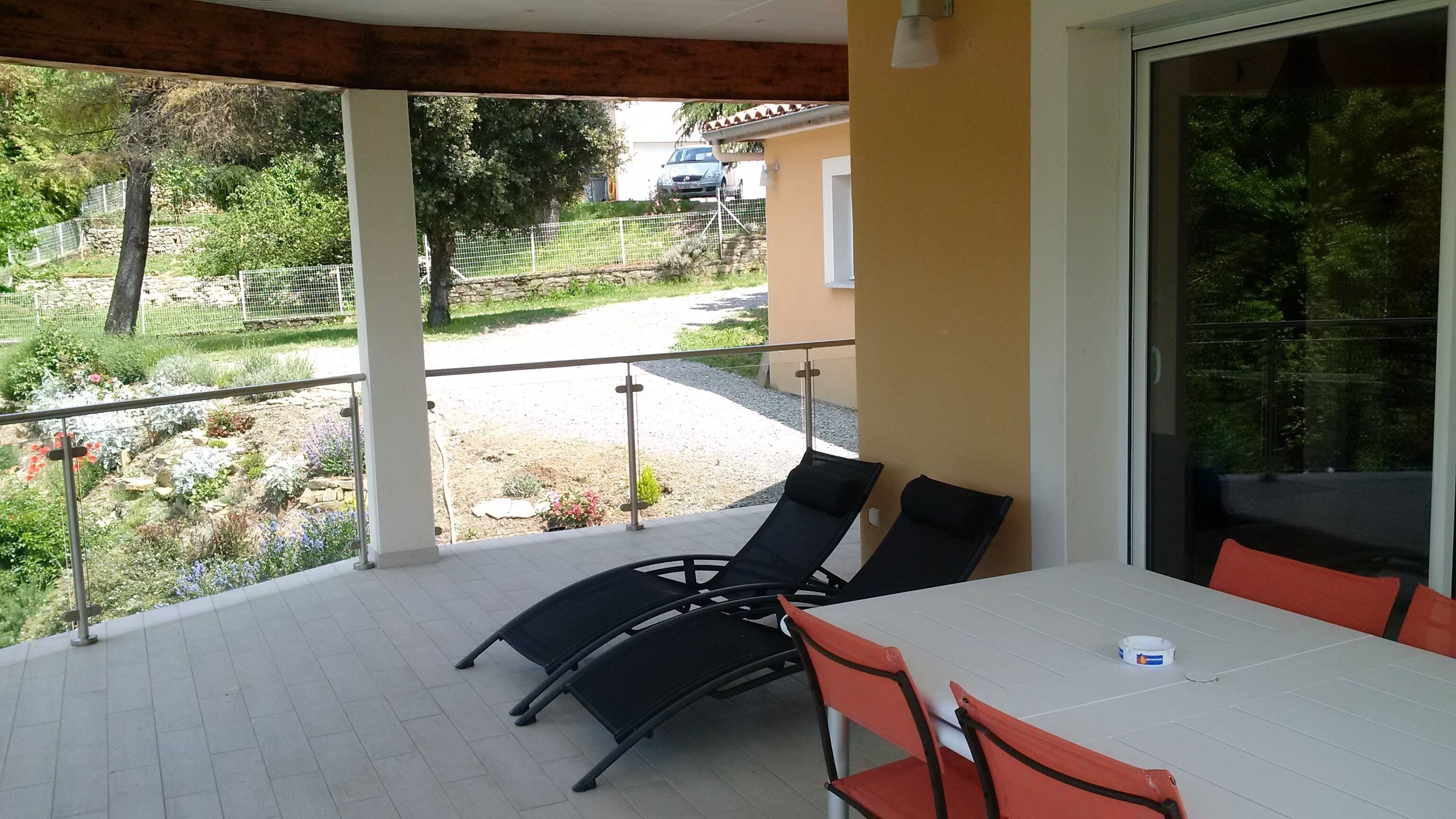 Eine überdachte Terrasse mit Tisch und Stühlen, eine Glastür sowie Blick auf einen Garten und ein geparktes Auto.