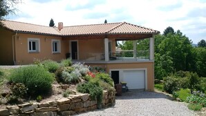 Uma casa de dois andares com varanda, garagem e um jardim com diversas plantas.