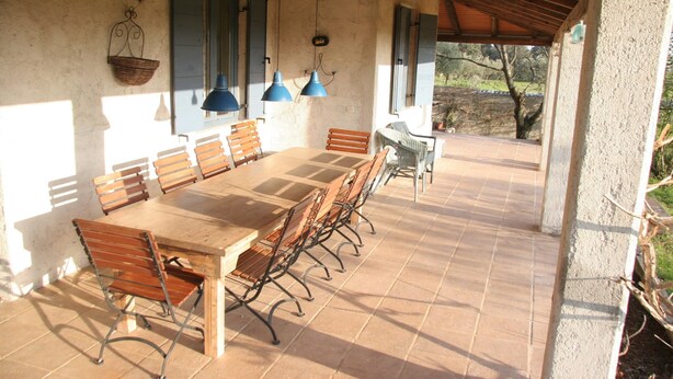 Een overdekt terras met een lange houten tafel en stoelen, blauwe hanglampen en uitzicht op het omliggende landschap.