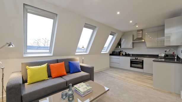 Un apartamento moderno con un sofá gris, una mesa de centro y una zona de cocina con armarios blancos y encimera negra.