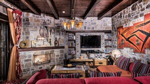 Une pièce chaleureuse avec un mur en pierre, un plafond en bois, une cheminée, un téléviseur et un coin salon confortable agrémenté de coussins rouges.