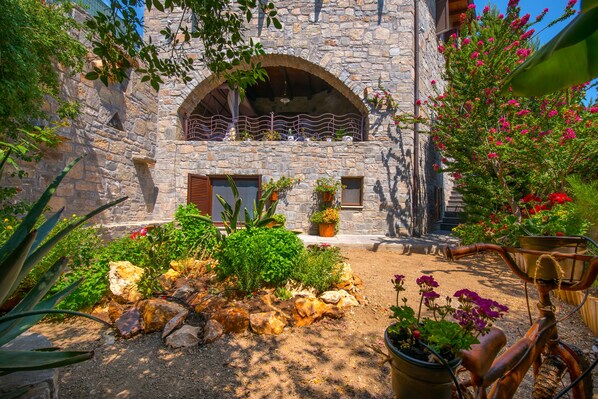 A stone building with an arched balcony, surrounded by a garden with various plants and a bicycle.