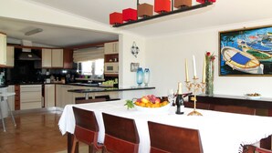 A dining area with a table set for four, a kitchen in the background, and a painting of boats on the wall. - Seazimbra - Luxury Apartment (Sesimbra)