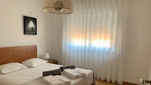 A neatly made bed with white linens and two rolled towels, a wooden headboard, a bedside lamp, and a framed picture on the wall. - Seazimbra - Luxury Apartment (Sesimbra)