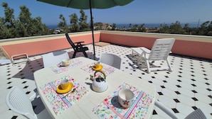 Eine Dachterrasse mit Tisch für zwei Personen, einem grünen Sonnenschirm und Blick aufs Meer.