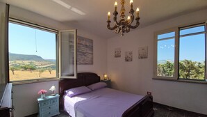 Ein Schlafzimmer mit einem Bett, einem Nachttisch, einer Kommode, einer Chandelieber und großen Fenstern mit Blick auf die Landschaft.