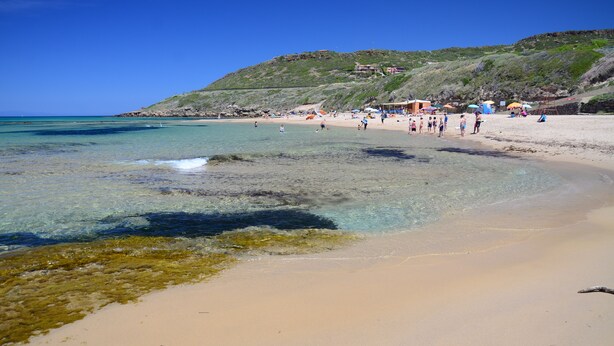 Ein Strand mit klarem Wasser, sandigem Ufer und einer Hügelkette im Hintergrund.