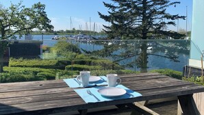 A wooden deck with a table set for two, overlooking a calm lake with boats and a tree in the background.