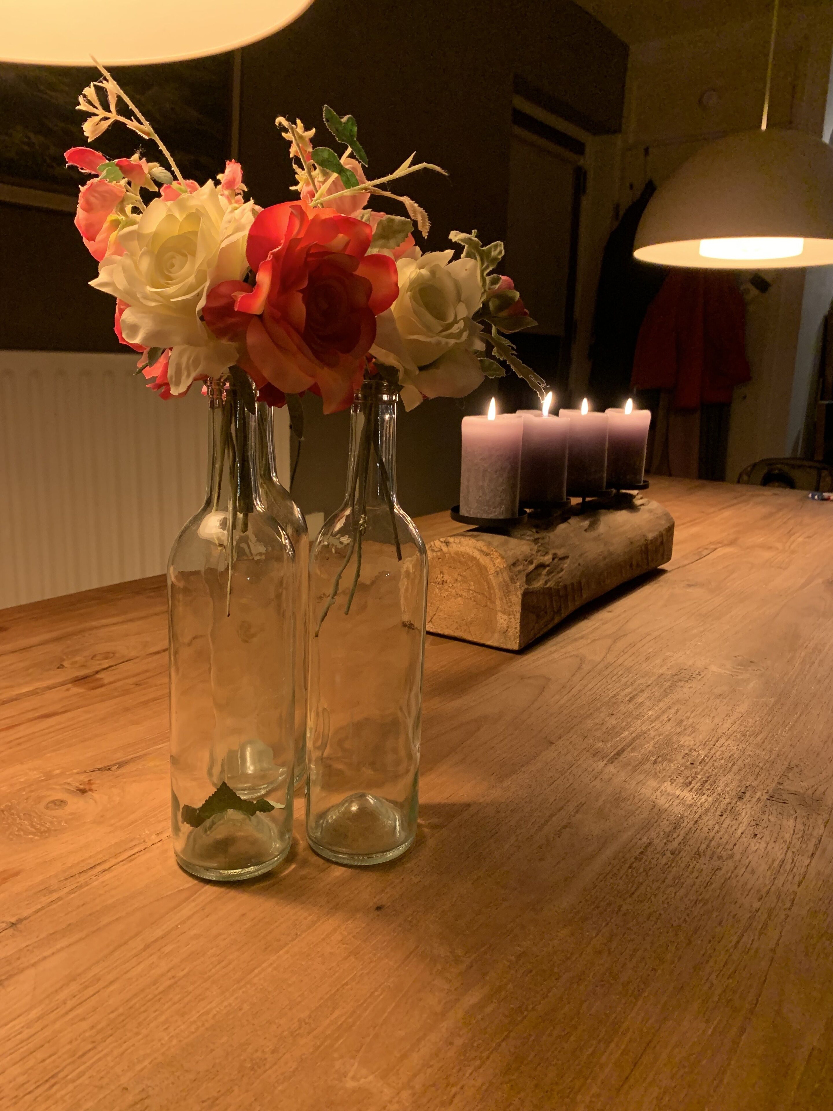 Twee heldere glazen vazen met bloemen op een houten tafel, een brandende kaars en een lamp op de achtergrond.