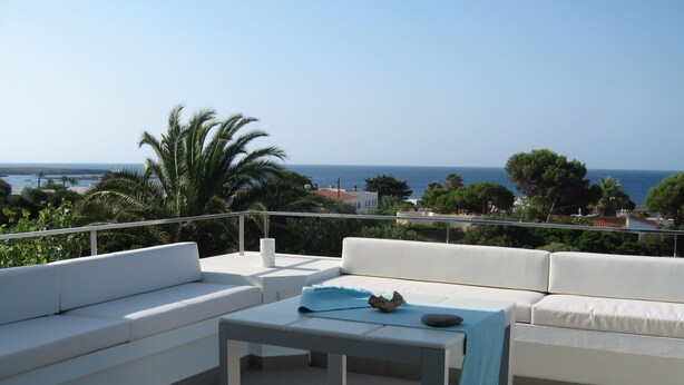 Een terras met witte sofa’s en een tafel, met uitzicht op de kust, palmbomen en de zee.