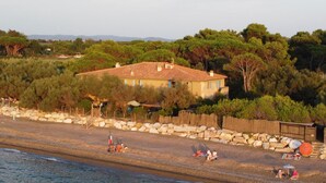 Ein Küsteneigentum mit einem großen Haus, einer Steinmauer und einem Strandabschnitt mit Menschen und Sonnenschirmen.