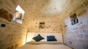 A stone-walled room with a bed, pillows, a small window, and a shelf with books.