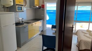 A compact kitchen with a microwave, sink, and a dining area with a chair. A bed with a view of the ocean is visible through an open door.