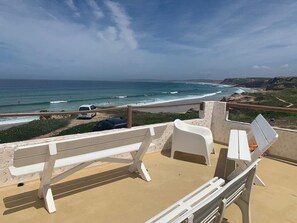 A beachside terrace with a white bench and lounge chairs, overlooking the ocean.