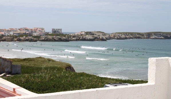 A coastal view with surfers in the water, a rocky shoreline, and buildings in the background.