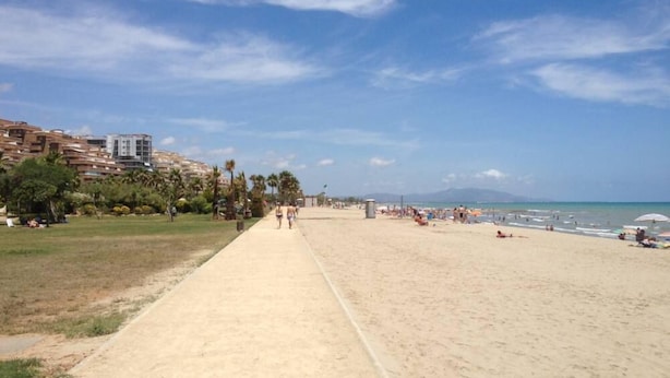 Une vaste plage de sable fin, avec un ciel dégagé, des palmiers et des bâtiments en arrière-plan.