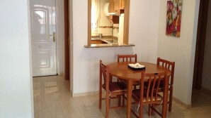 Un coin repas avec une table ronde en bois et quatre chaises, une cuisine visible à travers une fenêtre et une porte donnant sur une autre pièce.