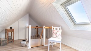 A high-ceilinged room with a sloped ceiling, a skylight, a wooden crib, a white chair, a small wooden table, and a woven basket. - Ferienwohnung Anton Greetsiel (Krummhoern)