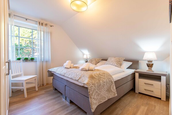 A bedroom with a bed, bedside tables, a chair, and a window with curtains. - Ferienwohnung Anton Greetsiel (Krummhoern)