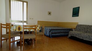 Ein Zimmer mit Holzboden, einem Bett mit gemusterten Textilien, einem Sofa, einem Esstisch mit Stühlen und einem Fenster mit Jalousien.