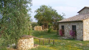 Un bâtiment en pierre avec un toit en tuiles, entouré d’une zone herbeuse parsemée de fleurs sauvages et agrémenté d’un arbre.