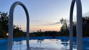 Une piscine dotée de deux échelles métalliques blanches, entourée d’arbres et surplombée d’un ciel dégagé.