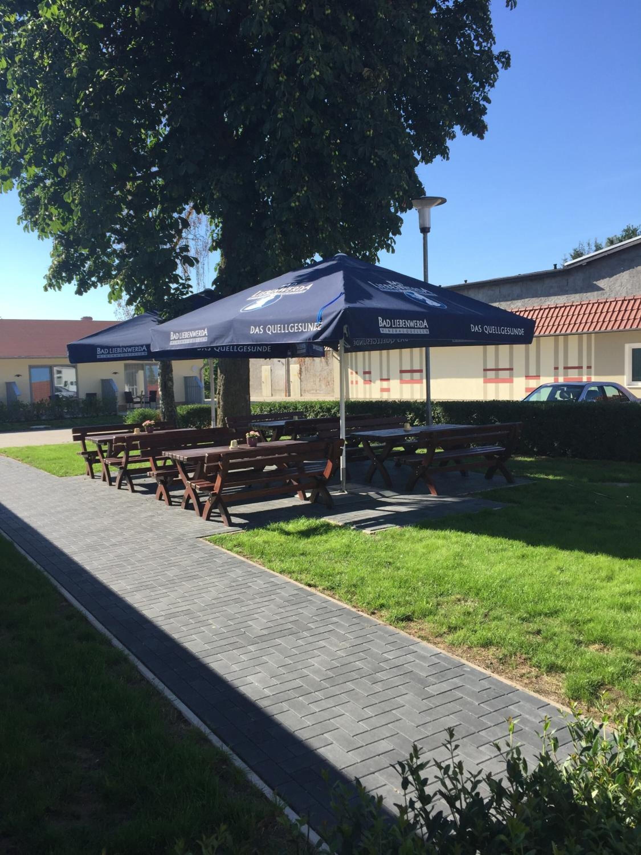 Un área de picnic con mesas y bancos bajo sombrillas azules, un sendero pavimentado y un edificio al fondo.