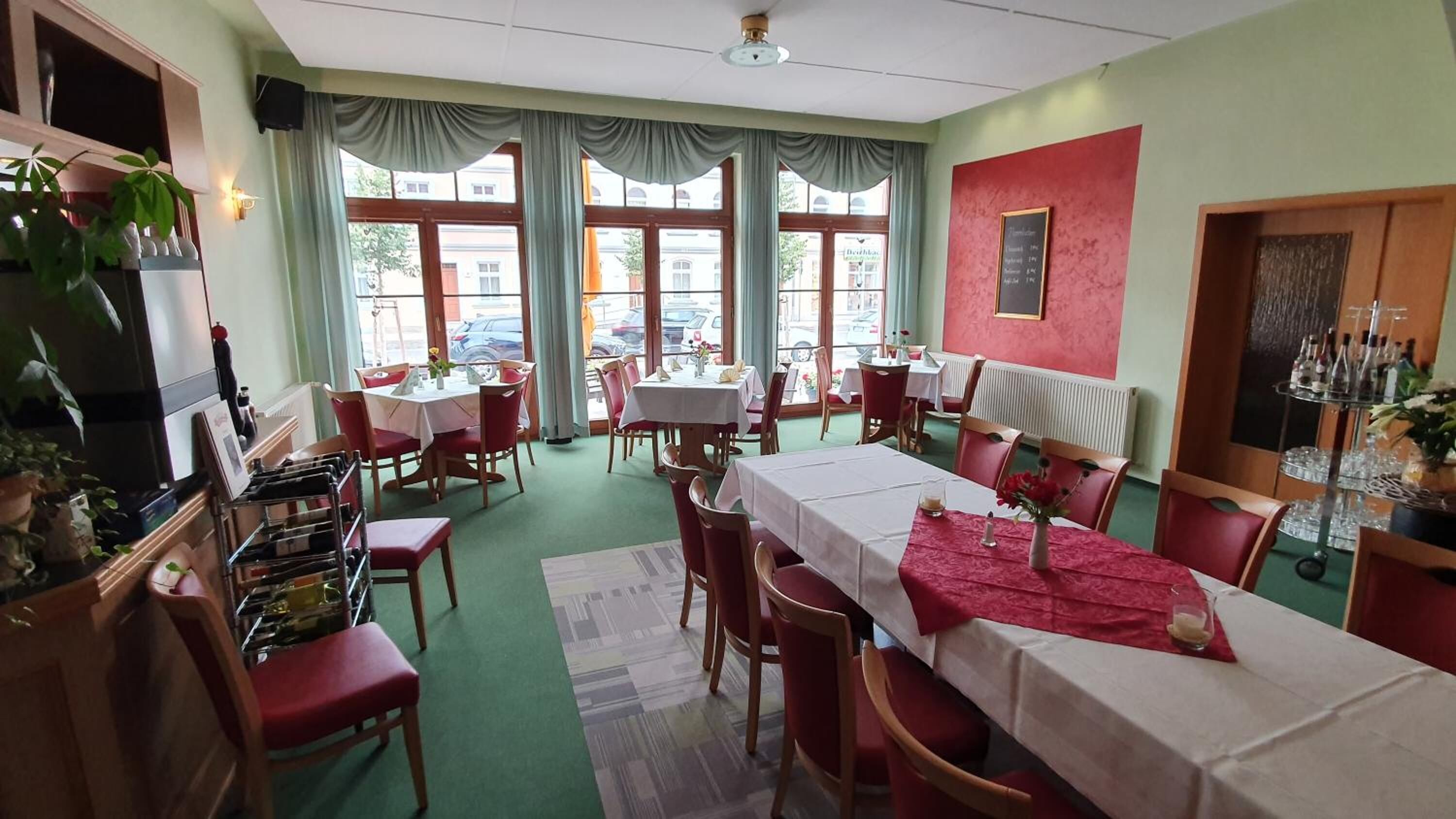 Uma área de refeições com mesas preparadas para quatro pessoas, grandes janelas com cortinas, uma parede vermelha com um cardápio emoldurado e uma planta à esquerda.