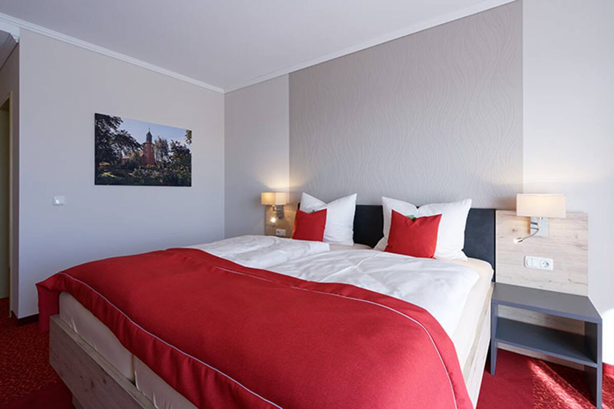 Habitación de hotel con una cama grande, ropa de cama roja, dos mesitas de noche con lámparas y un cuadro colgado en la pared.