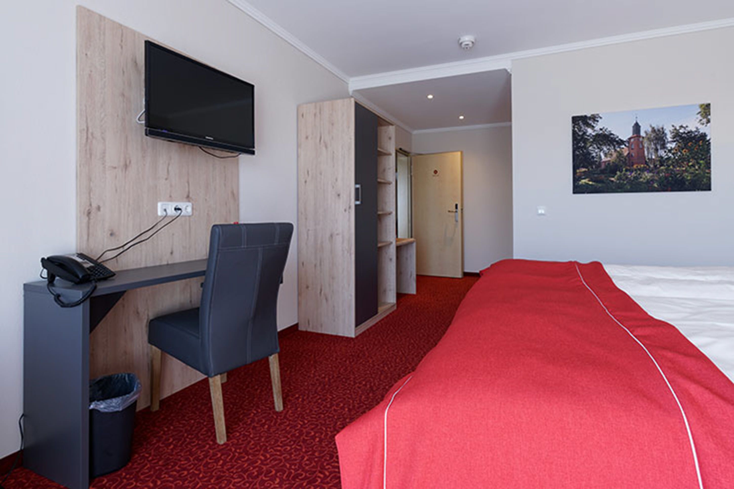 A hotel room with a desk, chair, TV, bed, and a red carpet.