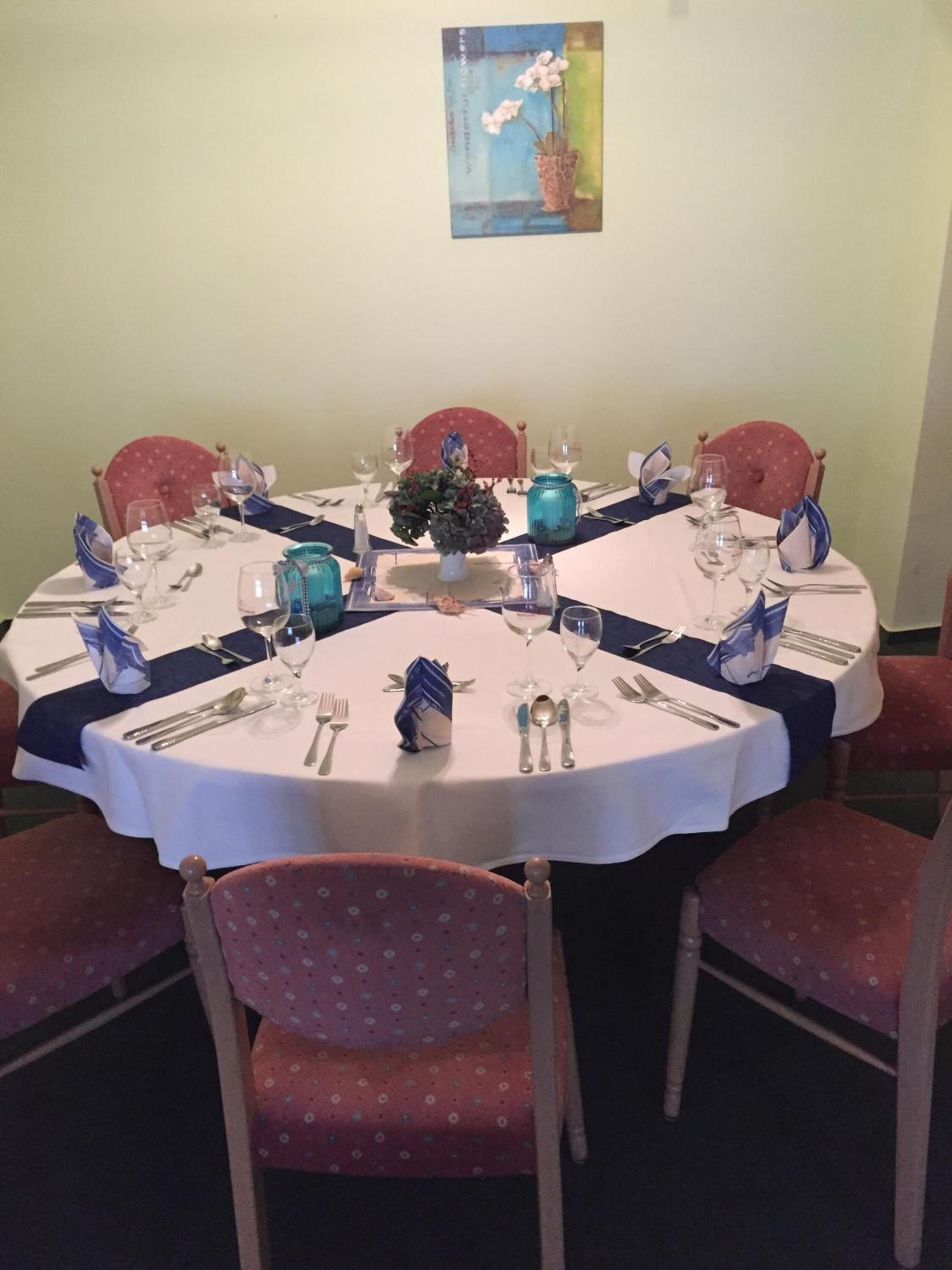 A round dining table set for six with white tablecloths, blue napkins, and matching glassware.