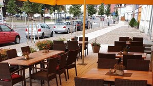 An outdoor café with wooden tables and wicker chairs under a yellow umbrella, set on a paved patio with potted plants. - Hotel Stadt Magdeburg (Perleberg)