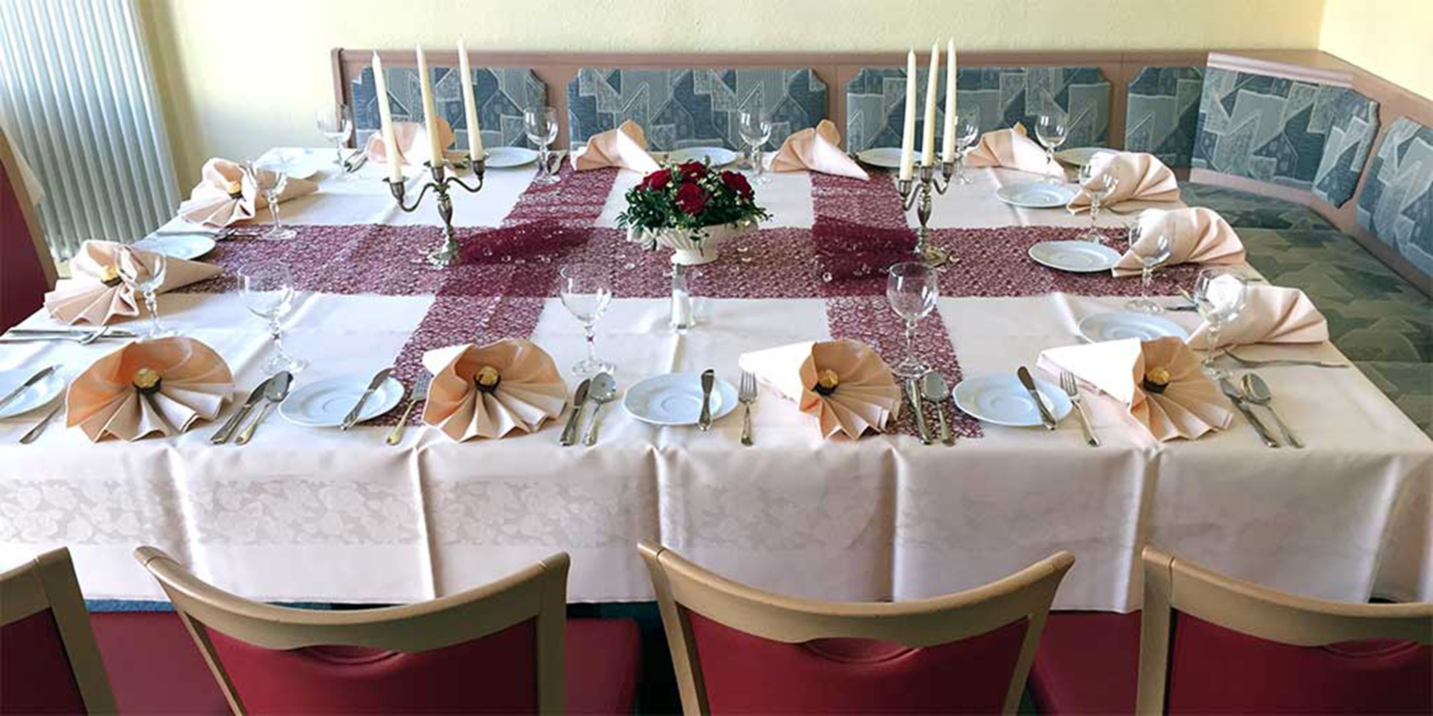 Mesa de jantar redonda posta para seis pessoas, com toalhas brancas, guardanapos vermelhos e um arranjo floral no centro.