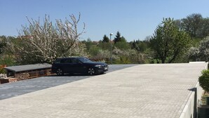 A paved parking area with a black car parked, surrounded by trees and a building with a sloped roof. - Hotel Stadt Magdeburg (Perleberg)