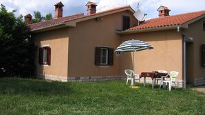 A house with a red-tiled roof, two chimneys, and a satellite dish. There is a table with chairs and a striped umbrella outside.