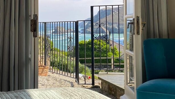 Ein Schlafzimmer mit großem Fenster und Vorhängen, einem türkisfarbenen Sessel und Blick auf das Meer und die Berge.