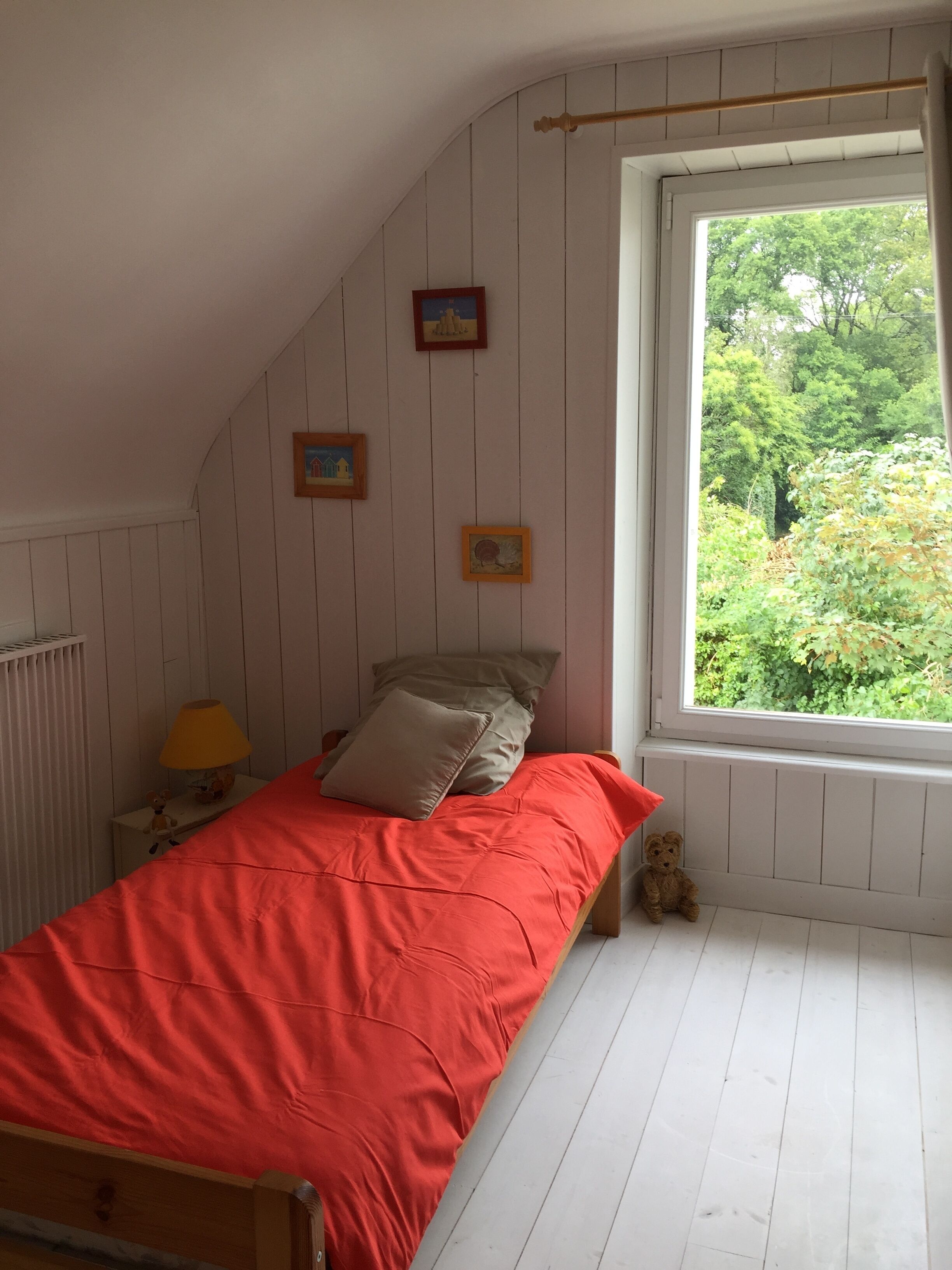 A bedroom with a sloped ceiling, a bed with a red bedspread, two pillows, a nightstand with a lamp, and a teddy bear. There are three framed pictures on the wall and a window with a view of greenery outside.