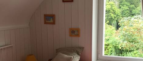 A bedroom with a sloped ceiling, a bed with a red bedspread, two pillows, a nightstand with a lamp, and a teddy bear. There are three framed pictures on the wall and a window with a view of greenery outside.