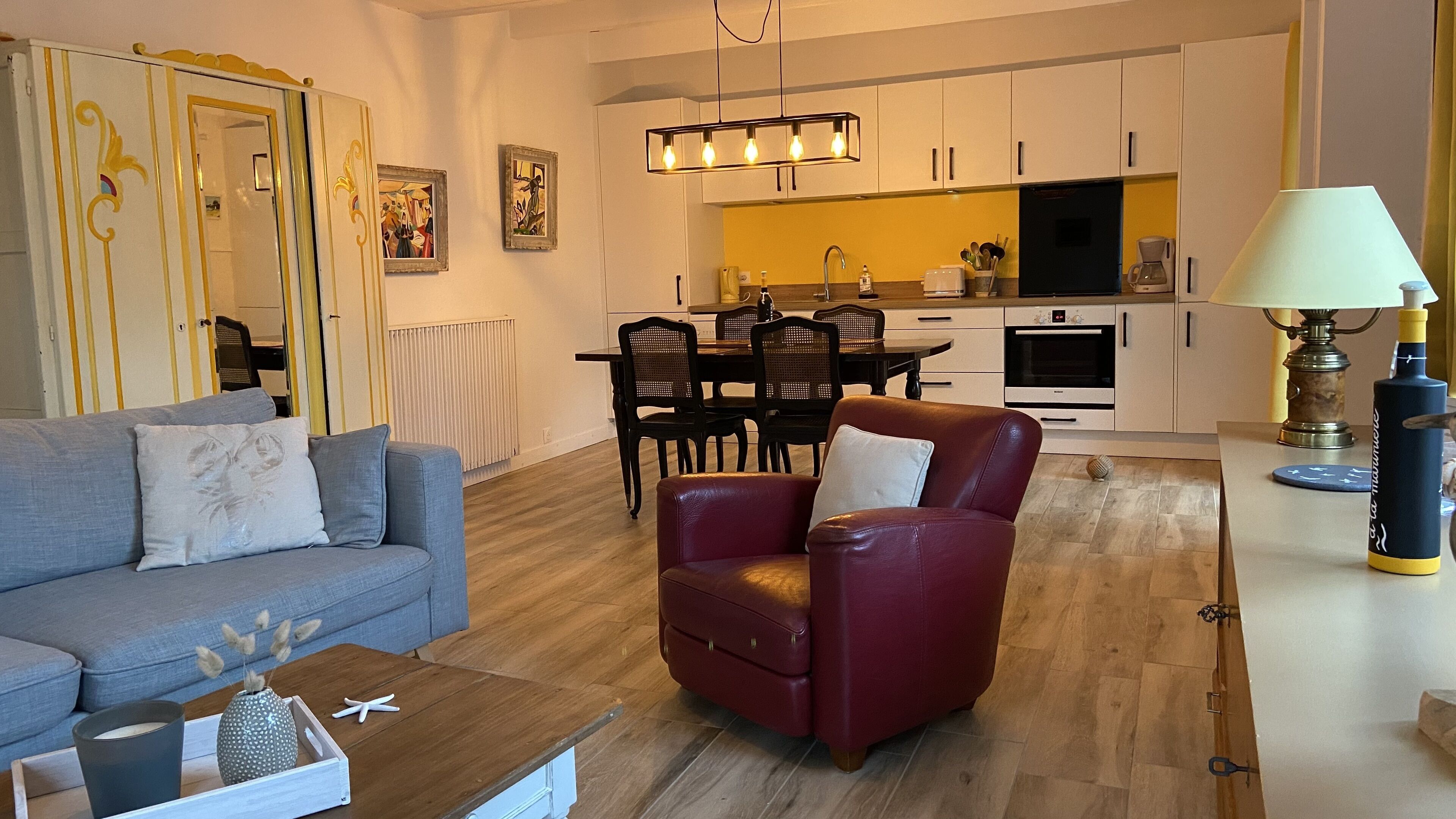 Una sala moderna con un sofá gris, un sillón rojo y una mesa de centro de madera. Hay una zona de comedor con una mesa y sillas, y una cocina con armarios blancos y un refrigerador negro.