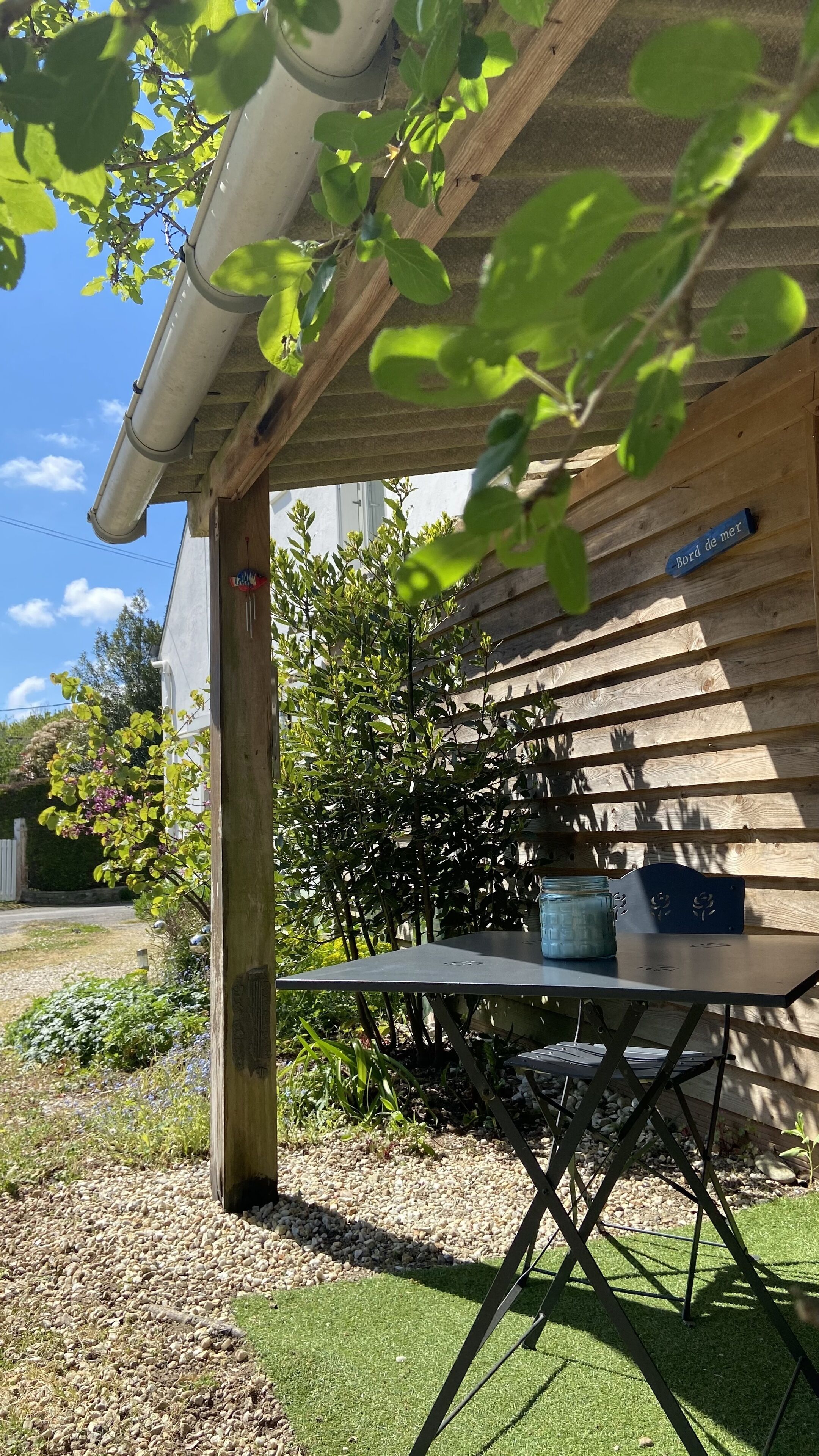 A wooden pergola with a table and chairs, a sign reading "Bord de mer", and a garden with gravel and grass.