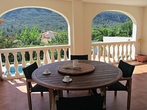A round wooden table with four chairs, a small table centerpiece, and a view of a mountainous landscape through arched windows.