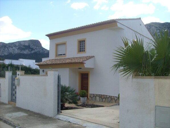 A two-story house with a tiled roof, a small front garden, and a mountain in the background.