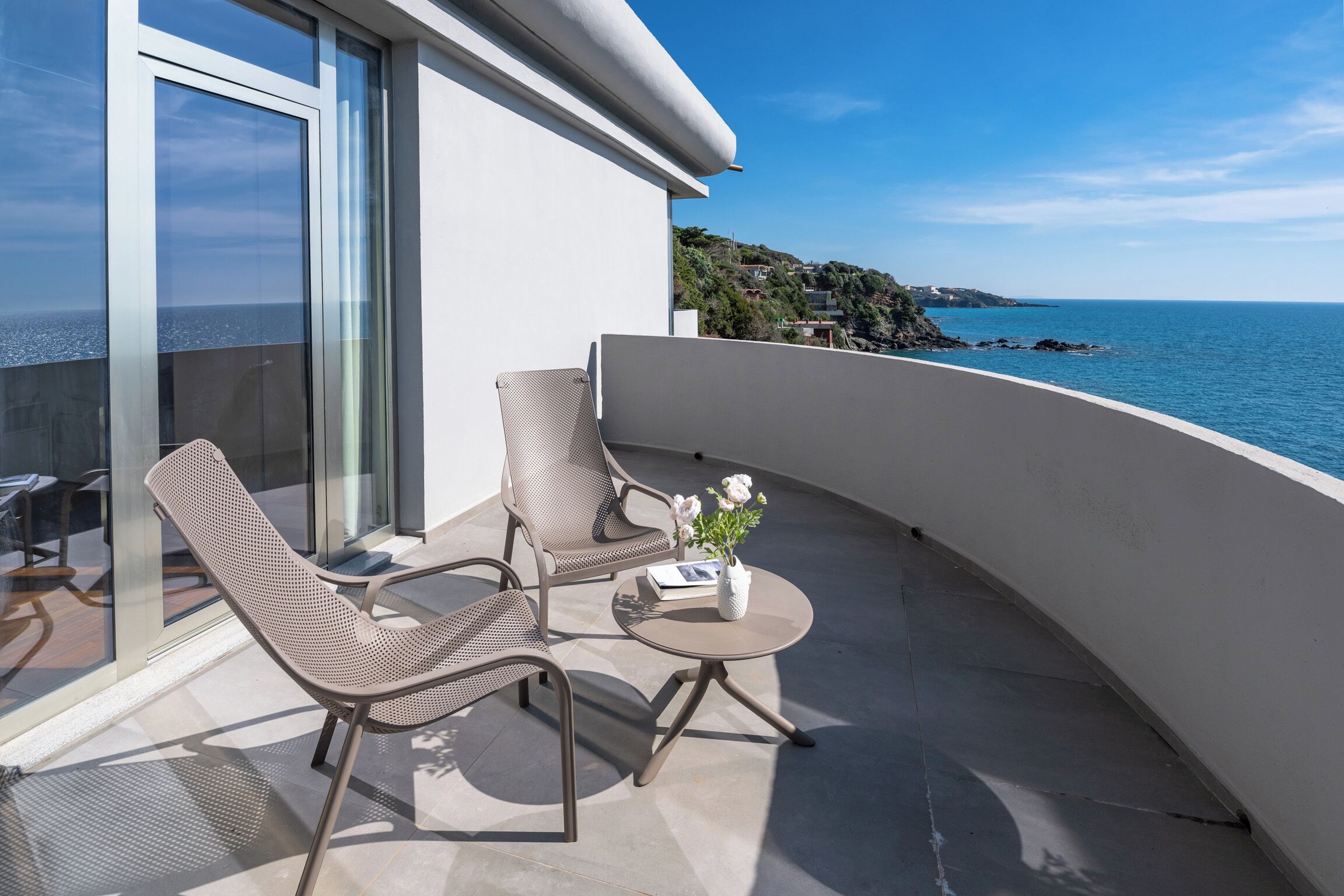 A balcony with two chairs and a small table, overlooking a coastal view.
