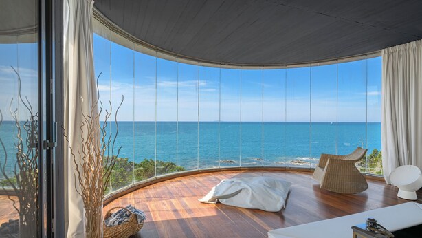 A room with a curved wooden floor, a large window with a view of the ocean, a white sofa, and a wicker chair. - House / Villa - Castiglioncello(LI) (Castiglioncello)